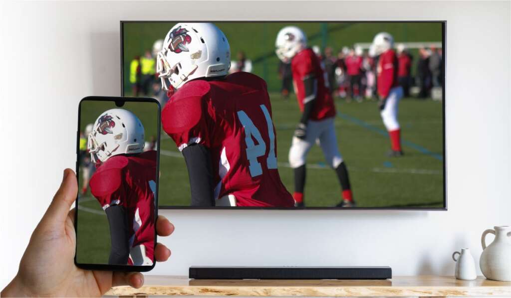 A hand holding a smartphone displaying an image of american footballers on the pitch. The same image is displayed on the wall-mounted Smart TV.